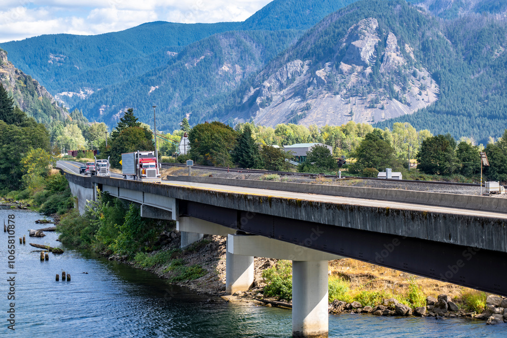 Fototapeta premium Red big rig classic semi truck transporting cargo in refrigerator semi trailer running in front of another semi trucks on the bridge across the tributary of the Columbia river in Columbia Gorge area