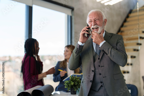 Elderly Caucasian man in a suit engages actively on a phone call, his experienced demeanor enlivened by the conversation, while colleagues confer in a modern office setting. photo