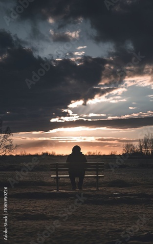 A solitary figure sits on a bench, silhouetted against a dramatic sky at sunset