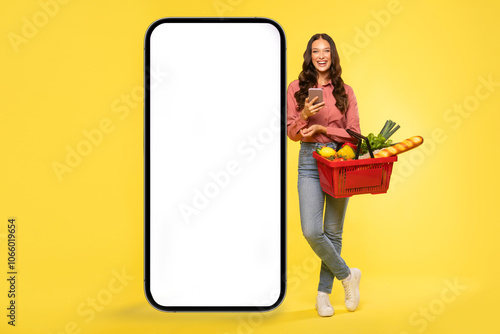 Happy woman with food products in basket posing near giant cellphone with white blank screen for mockup, isolated on yellow background. Delivery service from shop concept photo