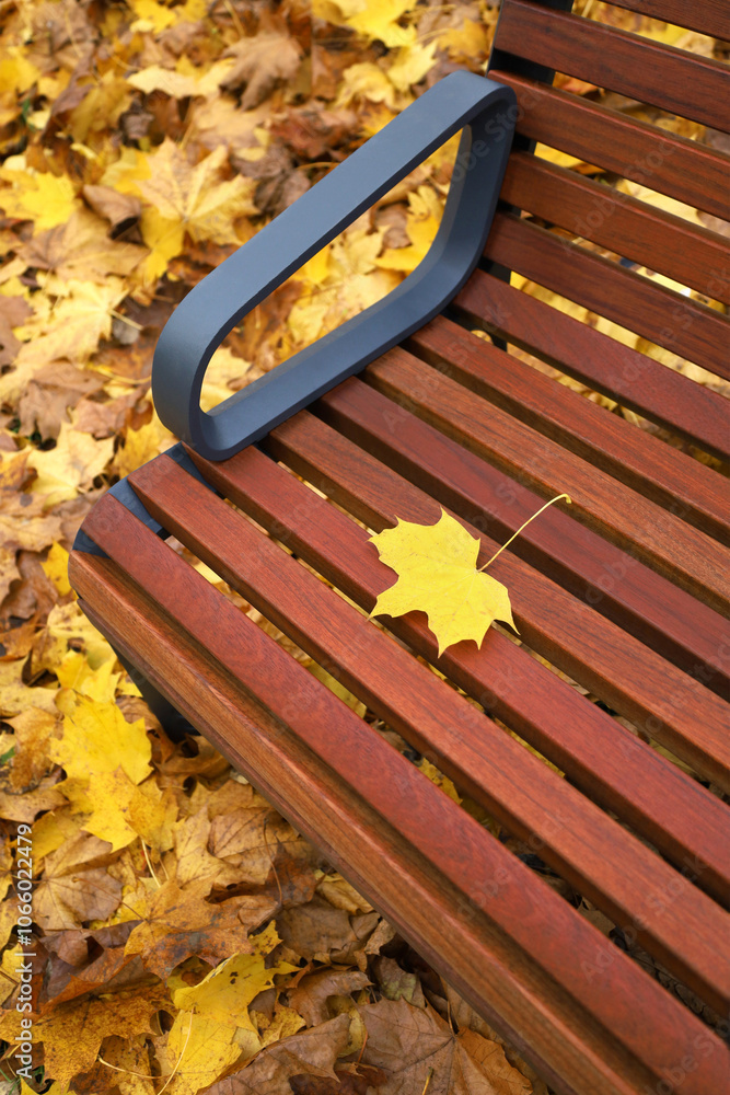 One small yellow maple leaf lies on brown wooden bench in autumn park. Fallen colorful leaves blurred on background. Fall mood vertical image.