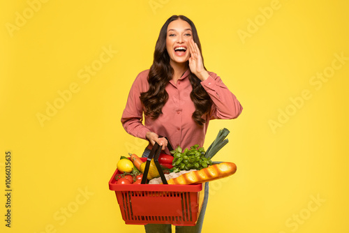 Young excited woman holding shopping basket full of food shouting with mouth wide open, announcing sales in supermarket, posing on yellow background photo