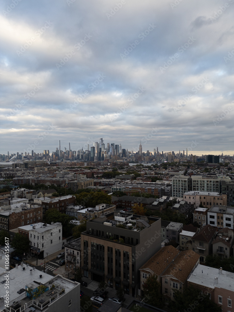 Wide aerial view capturing the neighborhoods of Hoboken and Jersey City, with the iconic New York City skyline stretching across the horizon in the background. Overcast skies add depth Wide aerial view capturing the neighborhoods of Hoboken and Jersey City, with the iconic New York City skyline stretching across the horizon in the background. Overcast skies add depth