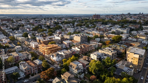 Wide aerial view capturing the neighborhoods of Hoboken and Jersey City, with the iconic New York City skyline stretching across the horizon in the background. Overcast skies add depth 