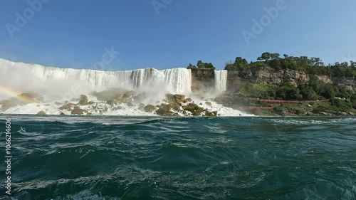 Force of Niagara waterfall cascades over rocks, creating mist and colorful rainbow under clear blue skies. Low-angle water surface moving point of view. Real time photo