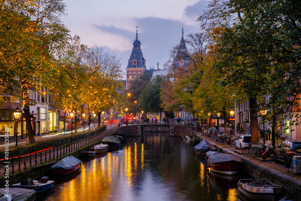 Fototapeta premium Golden leaves flutter above tranquil canals, reflecting the warm lights of bridges and buildings as twilight envelops Amsterdam.