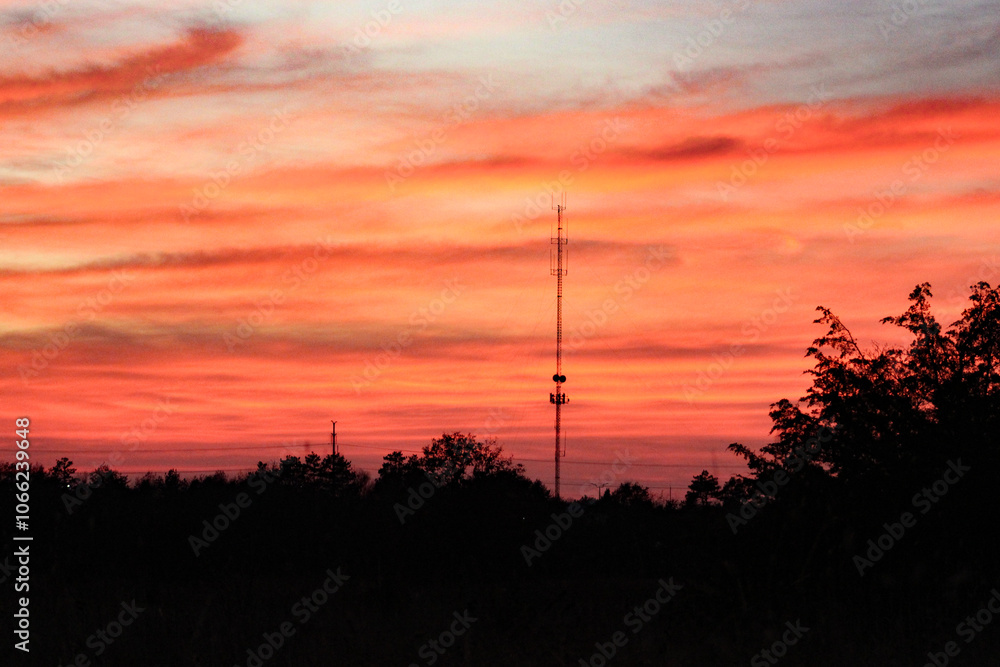 Obraz premium radio communication tower antenna against a vibrant sunset with tree silhouette news media broadcast area background