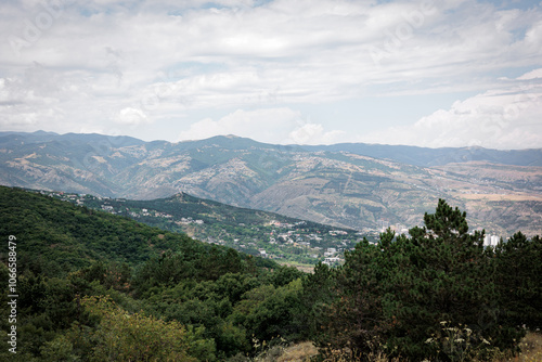 Summer mountain view of Tbilisi, Georgia. Soft clouds cast gentle light over the city and its green landscape, with lush trees in the foreground, creating a calm, scenic atmosphere