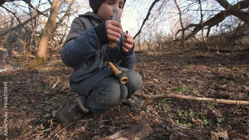 A young boy practices bushcraft skills in the forest, carefully carving a stick with a knife. This video captures outdoor learning and survival skills in a natural setting photo