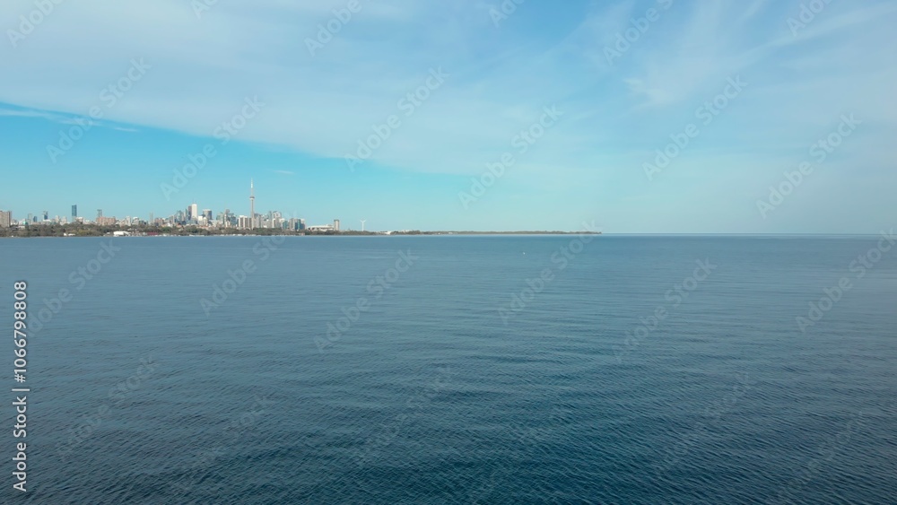 Fototapeta premium Toronto skyline rises peacefully over calm waters of Lake Ontario, blending modern architecture with serene natural beauty. Expansive lake and blue sky, harmony between city and nature in Canada