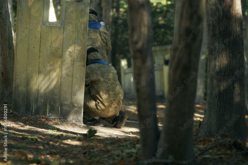 Paintball player in protective mask aims gun, takes cover under fire, paint splashes. Paintball player hide behind wooden barricade, shooting paintballs. Paintball players. Paintball player in protective mask aims gun, takes cover under fire, paint splashes. Paintball player hide behind wooden barricade, shooting paintballs. Paintball players.