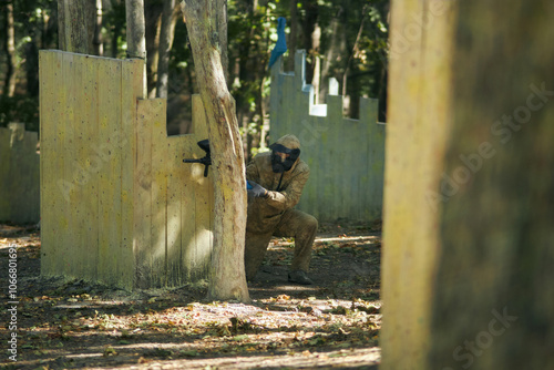 Paintball player in protective mask aims gun, takes cover under fire, paint splashes. Paintball player hide behind wooden barricade, shooting paintballs. Paintball players. photo