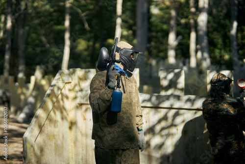 Paintball player in protective mask aims gun, takes cover under fire, paint splashes. Paintball player hide behind wooden barricade, shooting paintballs. Paintball players. photo