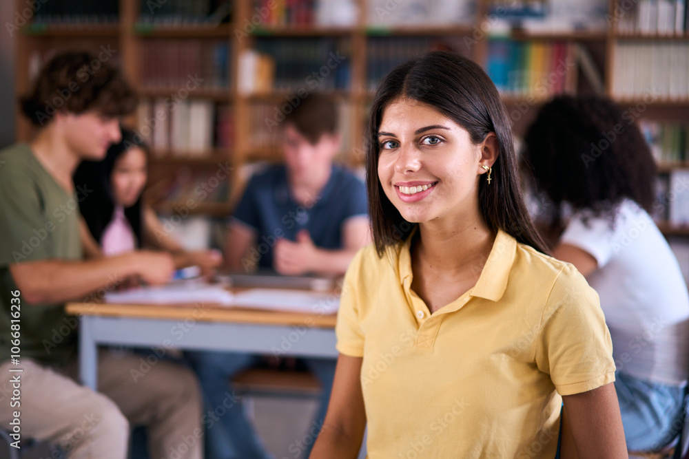 Portrait pretty young Caucasian female student sitting in library with classmates unfocused in background. Group friends meeting together in study room. High school girl poses looking happy at camera Portrait pretty young Caucasian female student sitting in library with classmates unfocused in background. Group friends meeting together in study room. High school girl poses looking happy at camera