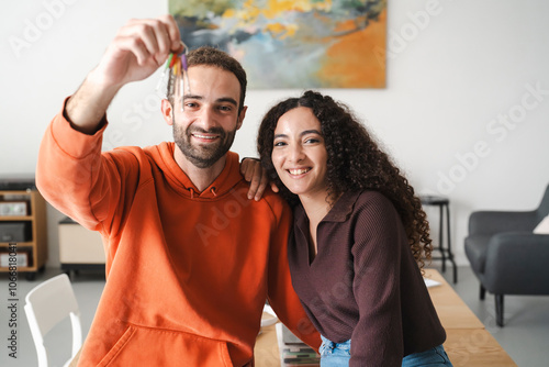 A cheerful young couple sitting together at a table, with the man in an orange hoodie holding up a set of keys, symbolizing a fresh beginning or new home in a brightly lit, modern room. photo