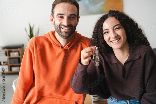 A smiling woman and man seated at a table, holding up keys, representing a new home or shared living experience, with modern art and plants in a bright, inviting living room. photo