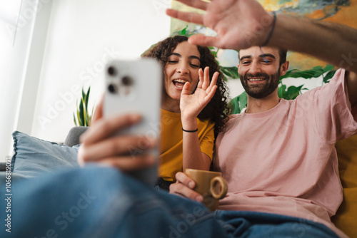 A cheerful couple seated on a couch, waving at a smartphone screen during a video call, with plants and cozy decor around them, highlighting a relaxed, homey environment. photo