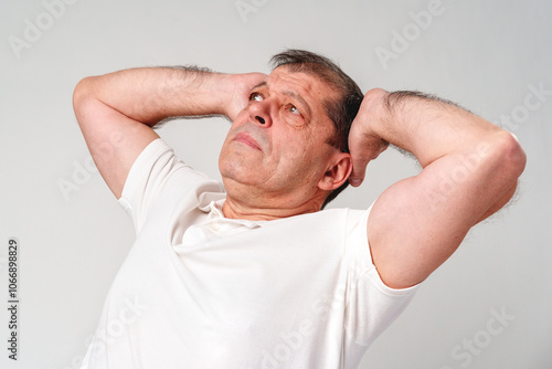 Elderly man stretching and relaxing indoors with a thoughtful expression and neutral background