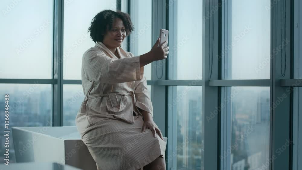 Medium shot of curvy young Biracial woman in soft light pink bathrobe sitting on elegant white freestanding bathtub in luxurious high-rise bathroom with city view, taking selfie portrait on smartphone Medium shot of curvy young Biracial woman in soft light pink bathrobe sitting on elegant white freestanding bathtub in luxurious high-rise bathroom with city view, taking selfie portrait on smartphone