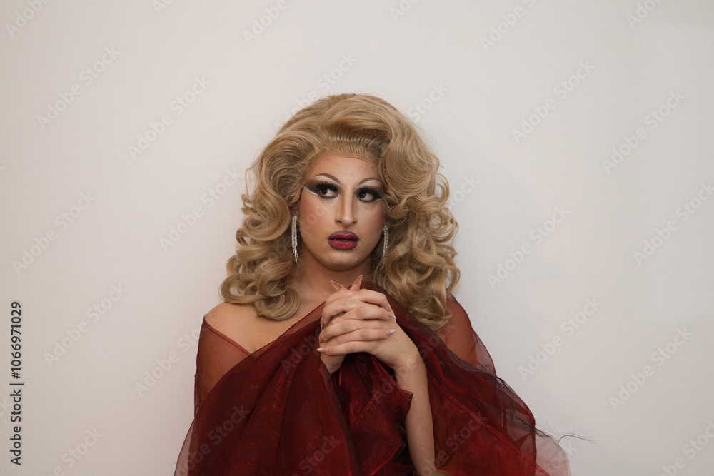 Fototapeta premium A drag queen with blonde wig and luxurious earrings and covered in red silk and on a white background with clasped hands praying imitating the virgin. Concept of lgtbqi rights and equality.