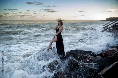 Quadro em tela A beautiful young girl in a long black dress stands on large stones on the shore of a raging ocean