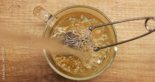 Person pours boiling water into glass mug with metal tea filter clip infuser spoon. Process of brewing herbal beverage on wooden board countertop