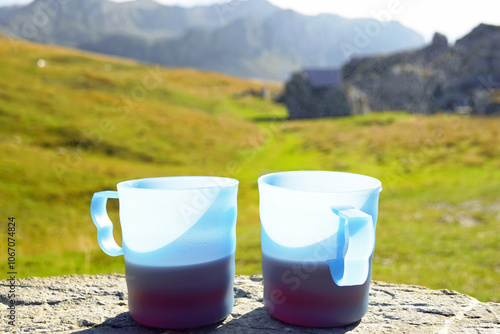 Picnic in a mountain valley: two plastic hiking mugs with black tea standing against the backdrop of bright green grass and beautiful mountain slopes. A rest stop during a hike in Lukavica, Montenegro photo