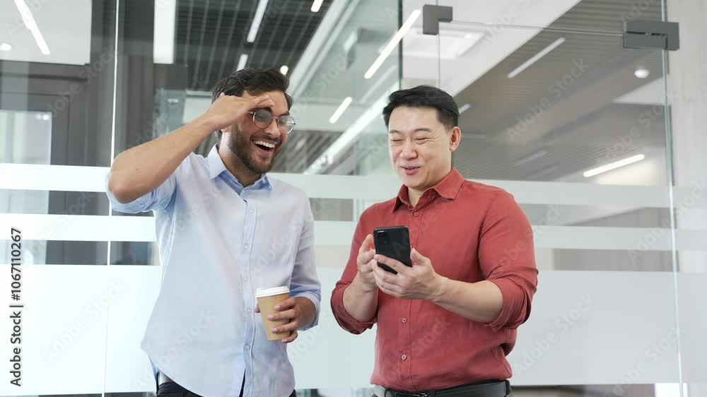 Two coworkers laughing and chatting over a phone in a modern business office. Happy cheerful team enjoying friendly conversation, fostering teamwork and workplace camaraderie during a casual moment