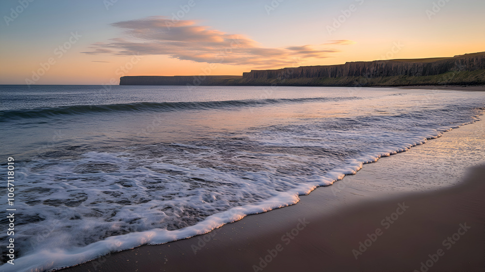 Obraz premium A serene beach scene at sunrise, featuring a calm ocean with gentle waves lapping at the shore and a dramatic cliff face in the distance.