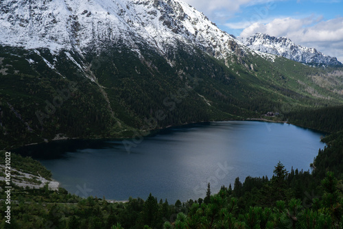 Fototapeta Naklejka Na Ścianę i Meble -  Morskie Oko Snowy Mountain Hut in Polish Tatry mountains, drone view, Zakopane, Poland. Aerial view shot of beautiful green hills and mountains in dark clouds and reflection on the lake Morskie Oko