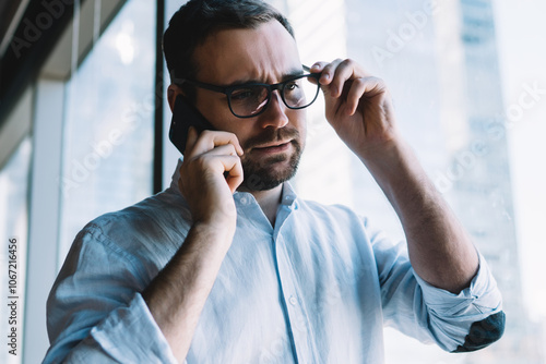 Caucasian employer in classic spectacles for provide eyes protection using cellphone gadget for making service consultation during work day, puzzled businessman calling to partner via cellular app