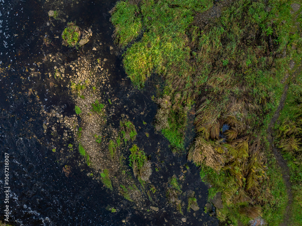 Top-down aerial photo of the Jagala River near Jägala Waterfall in Estonia. On the left the river flows with dark, almost black water speckled with white foam. On the right rugged green landscape. Top-down aerial photo of the Jagala River near Jägala Waterfall in Estonia. On the left the river flows with dark, almost black water speckled with white foam. On the right rugged green landscape.
