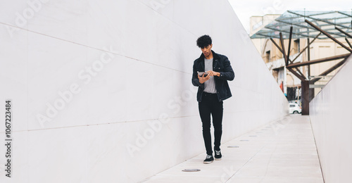 Stylish Latino hipster guy updating profile in social networks while walking at city urban setting and using 4g wireless for browsing, trendy man blogging for communicate with virtual friends