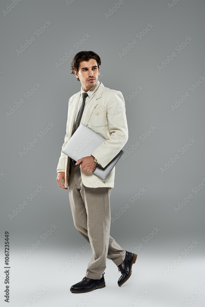A young man dressed in elegant formal wear stands gracefully, holding a laptop amidst a modern studio setting. A young man dressed in elegant formal wear stands gracefully, holding a laptop amidst a modern studio setting.