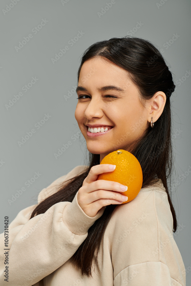 A stylish young woman beams with happiness, holding an orange close to her cheek. A stylish young woman beams with happiness, holding an orange close to her cheek.