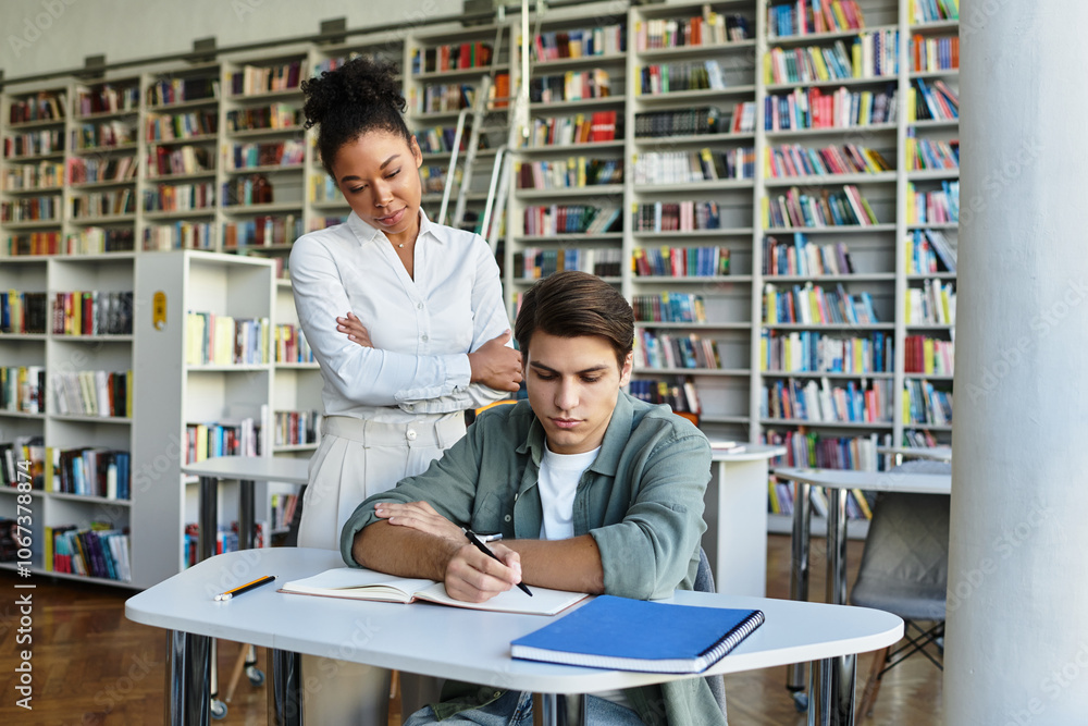 A student diligently writes in his notebook as a teacher watches with interest, surrounded by books. A student diligently writes in his notebook as a teacher watches with interest, surrounded by books.