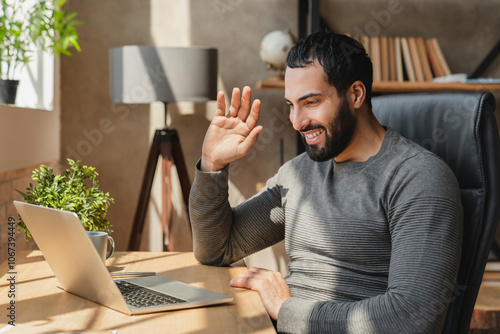 Distant communication concept. Smiling arab man waving hand at laptop camera, making video call. Happy guy chatting with family or having online meeting, sitting on chair at home photo