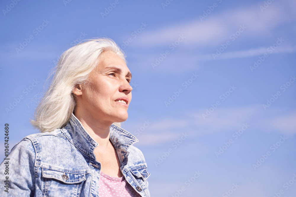 Confident senior woman with white hair in denim jacket enjoying sunny day against blue sky background. Confident senior woman with white hair in denim jacket enjoying sunny day against blue sky background.