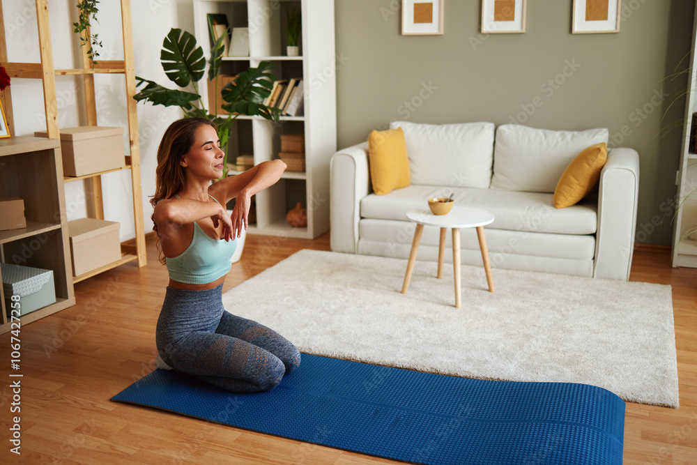 Fototapeta premium Young woman stretching arms while kneeling on yoga mat at home