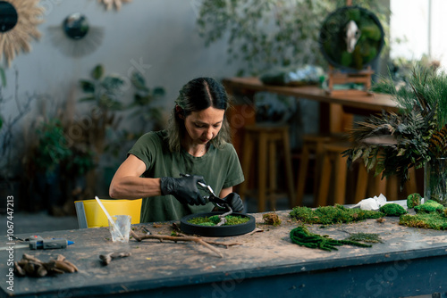 in large green workshop a thin woman with green hair in black gloves at a large table collects a composition from stabilized moss photo