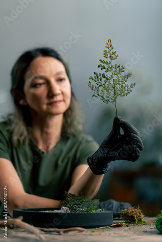 Close-up in a large green workshop, a woman with green hair in black gloves holds a sprig of green living plant nature protection decor element