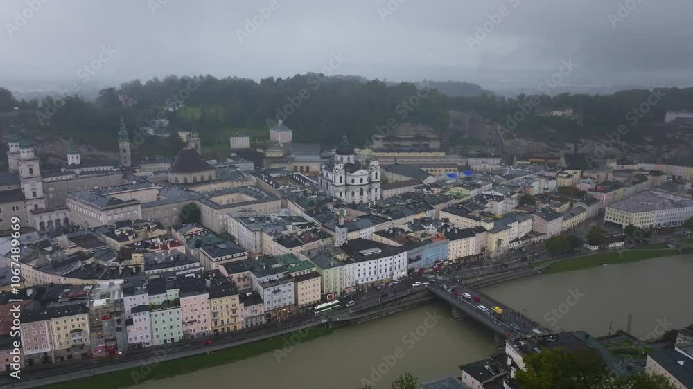 Aerial views highlight Salzburg, Austria, as the Salzach River meanders through the city. Panning reveals a mix of historic buildings and modern charm beneath a cloudy sky Aerial views highlight Salzburg, Austria, as the Salzach River meanders through the city. Panning reveals a mix of historic buildings and modern charm beneath a cloudy sky