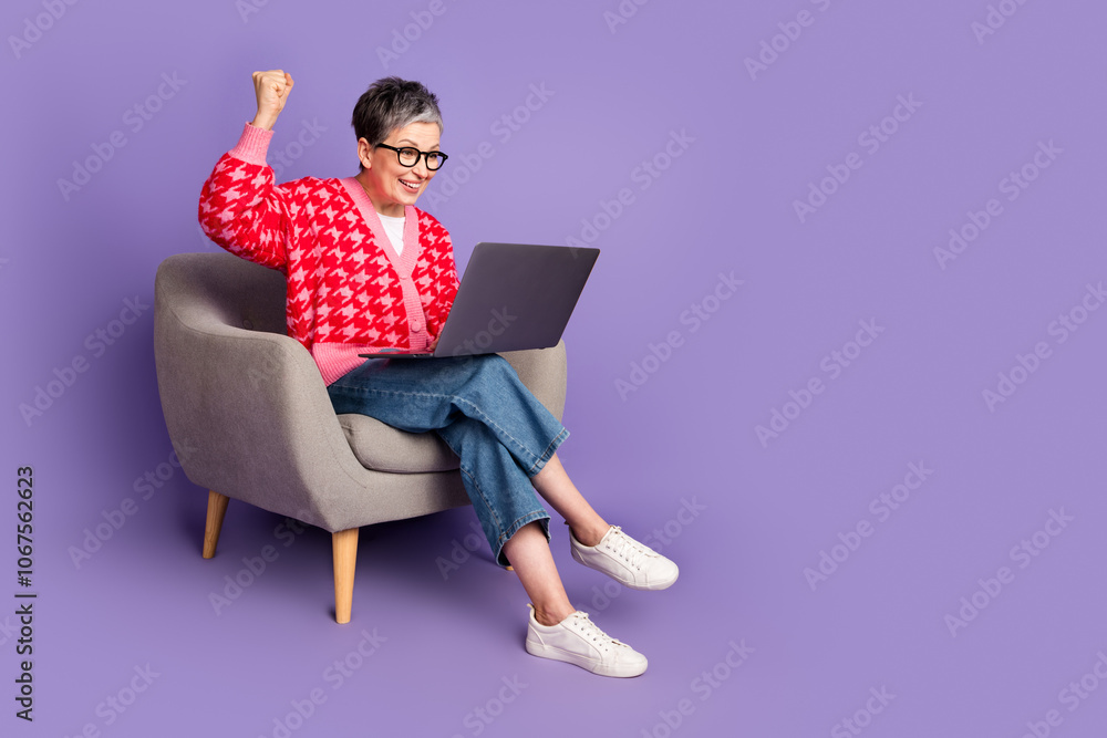 Full size photo of pretty retired female sit armchair hold netbook winning wear trendy red outfit isolated on purple color background Full size photo of pretty retired female sit armchair hold netbook winning wear trendy red outfit isolated on purple color background