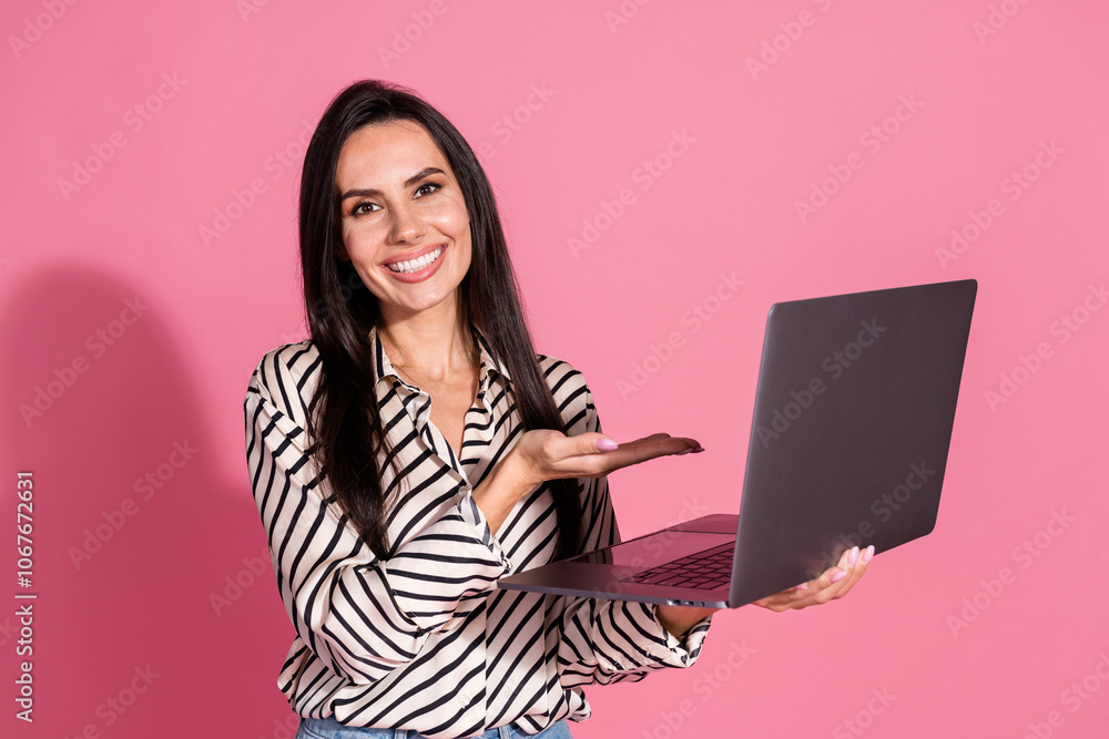 Naklejka premium Cheerful young woman with brunette hair promoting technology against pink background holding laptop in striped shirt pointing at screen