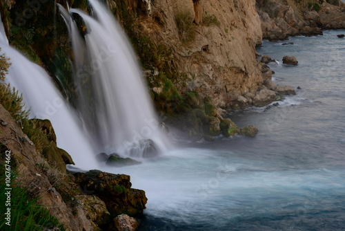 Scenic view of Duden Waterfall in Antalya, Turkey, cascading from cliffs into the Mediterranean Sea with a faint rainbow visible. 