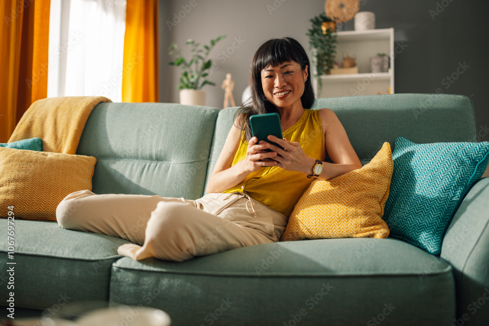 Obraz premium Asian woman relaxing on green couch while using smartphone, bright room