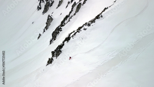 single snowboarder glides across a steep, open face of pure snow, framed by dark rocky outcrops.