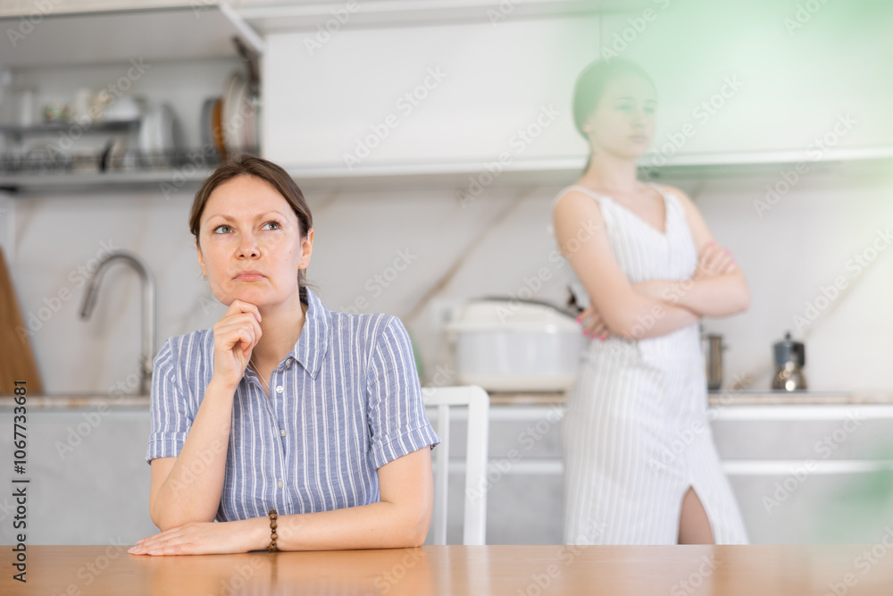 Angry mother sitting at the table turning her back upon offended teenage daughter standing at the kitchen cupboard Angry mother sitting at the table turning her back upon offended teenage daughter standing at the kitchen cupboard