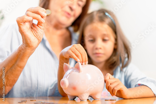 Grandmother and granddaughter inserting coins into piggy bank together, creating heartwarming moment of bonding and teaching value of saving at home 