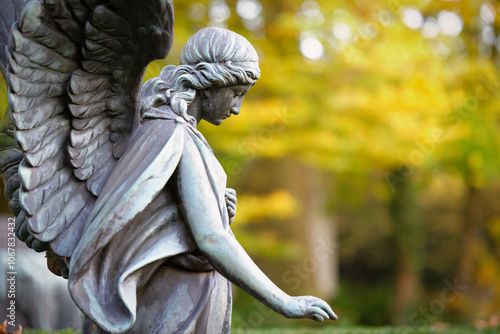 a winged angel figure in profile in an autumnal cemetery looks sadly to the ground with an outstretched hand